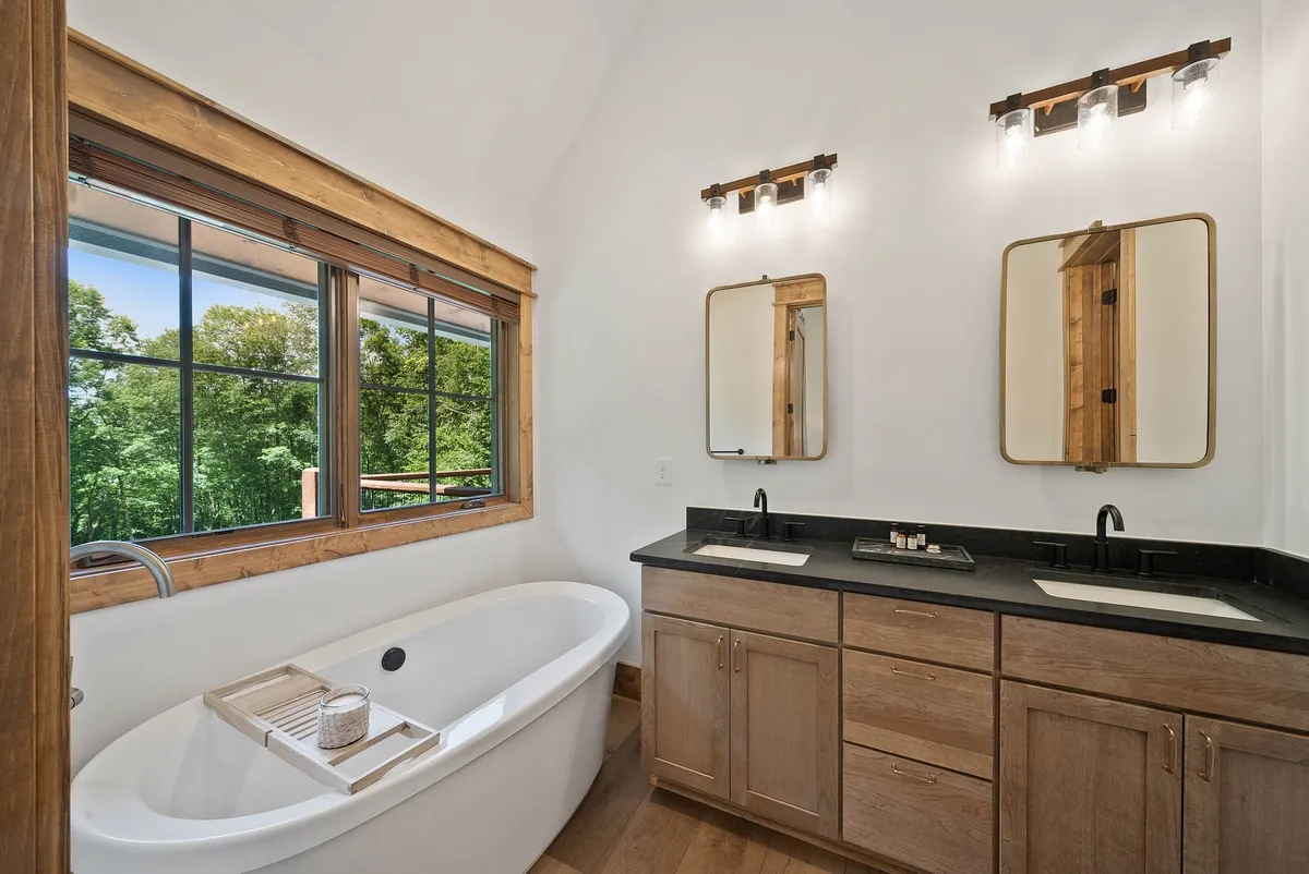 A bright and modern bathroom with a freestanding bathtub, a double sink vanity, and large windows offering views of the outdoors. The room is bathed in natural light, creating a spa-like, rejuvenating environment.