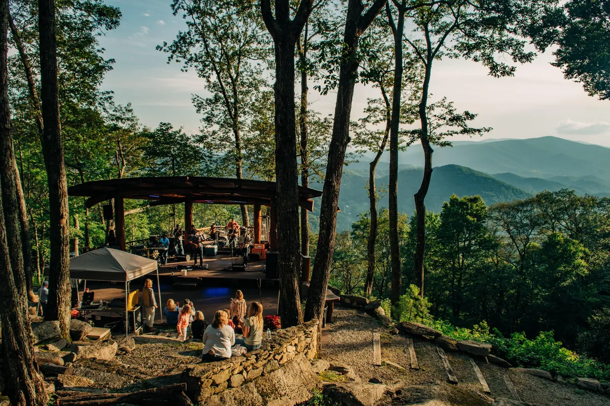 An outdoor wooden stage set against a mountain backdrop, with a gathering of people enjoying a live music performance. The natural stone pathway, picnic setup, and joyful crowd evoke a festive, community-oriented atmosphere.