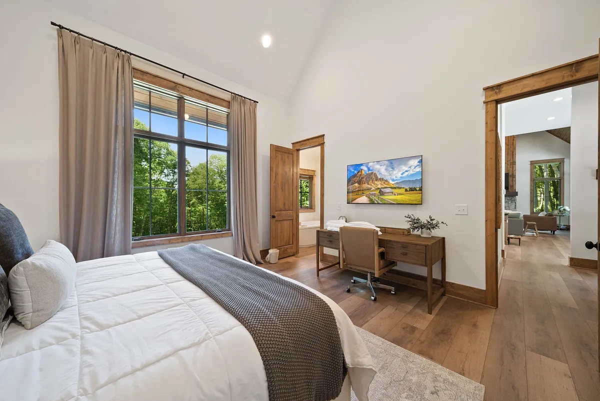 A tranquil bedroom scene with a textured bed, a work desk and chair, a television mounted on the wall, and a large window overlooking lush greenery. The space is filled with natural light and decorated in calming, earthy tones.