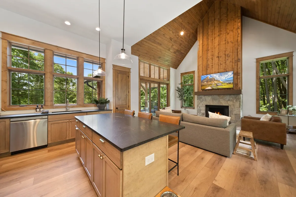 An open-concept kitchen and living area with a central island, a crackling fireplace, and expansive windows. The room exudes warmth with its wood finishes and offers plenty of space for preparing meals, entertaining, or simply relaxing by the fire.