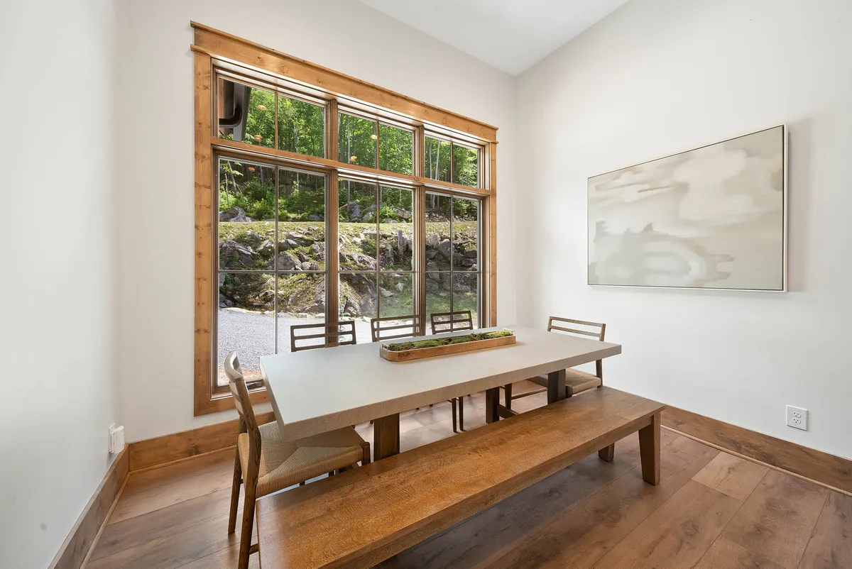 A serene dining area centered around a wooden table, surrounded by large windows that offer picturesque views of the surrounding woods. The minimalist décor and abundant natural light create a tranquil, welcoming space for shared meals and conversation.