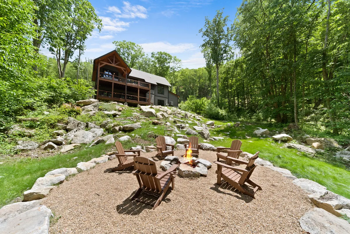 A spacious outdoor area featuring a fire pit surrounded by Adirondack chairs and lush greenery, with a two-story cabin in the background. The scene suggests a welcoming space for family bonding, storytelling, and evening gatherings under the stars.