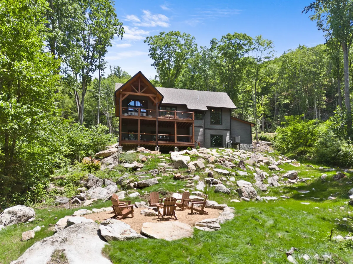 The image showcases a rustic yet luxurious mountain cabin nestled among lush greenery and rocky outcrops. A spacious deck overlooks the scenic surroundings, while a cozy fire pit area invites guests to gather and unwind. The natural landscape creates a peaceful setting, hinting at outdoor adventures and tranquil relaxation just steps from the cabin.