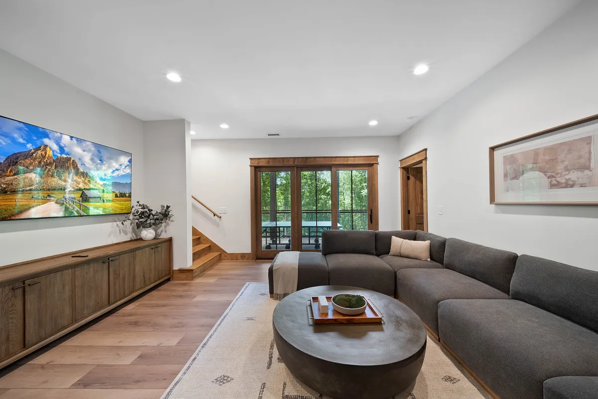 A spacious entertainment room featuring a large sectional sofa centered around a round coffee table, with a smart TV mounted across from the seating area. Expansive windows offer natural light and views of the outdoors, creating a comfortable, modern space for relaxing and socializing.