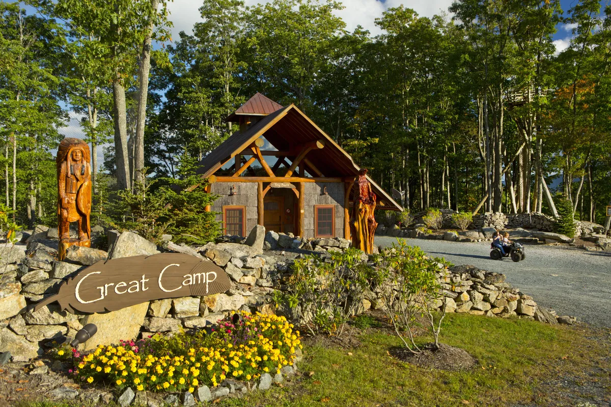 A rustic wooden lodge with carved totem poles, a colorful flower bed, and signage reading 'Great Camp.' The setting is inviting and designed for exploration, socializing, and outdoor fun amid nature trails and gathering spots.