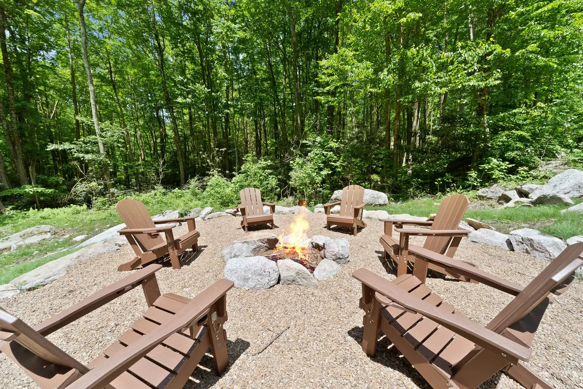 A cozy fire pit surrounded by Adirondack chairs set in a forested area, perfect for gathering, storytelling, and stargazing. The mood is warm and social, inviting intimate connection with nature and each other.