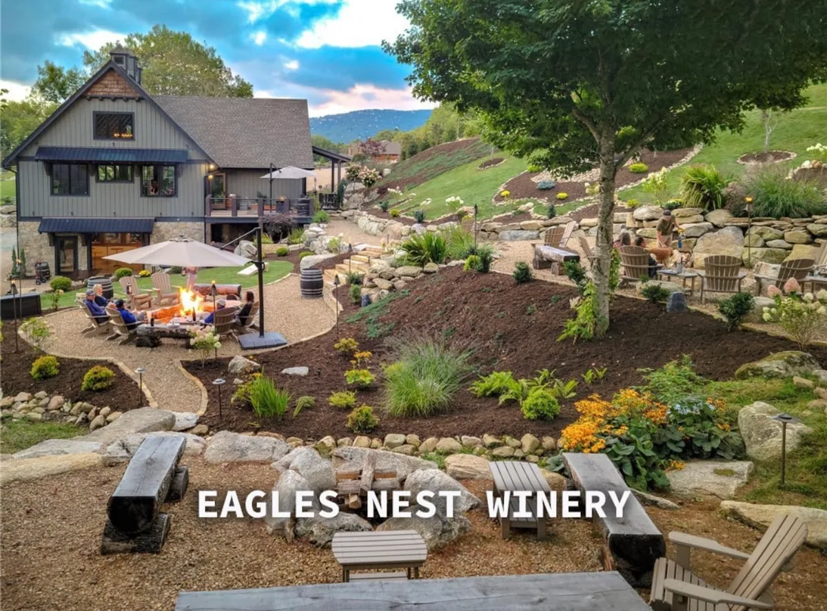 This image showcases a welcoming fire pit with ample seating, a view of the vineyard, landscaped gardens, and wine barrels. It captures people socializing, relaxing by the fire, and enjoying scenic views in a tranquil, upscale outdoor setting.