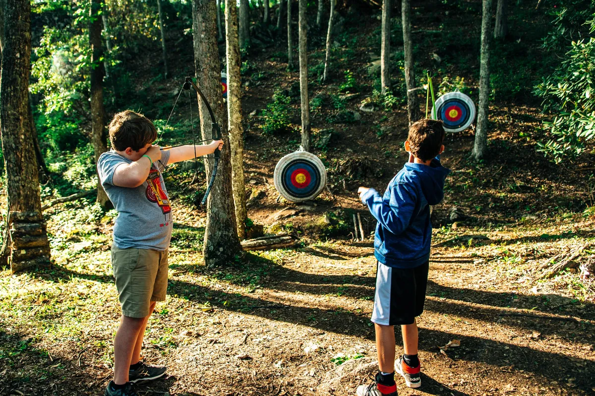Children practice archery outdoors, aiming at colorful target boards set among tall trees. The atmosphere is lively and engaging, with a clear focus on learning new skills and enjoying nature together.