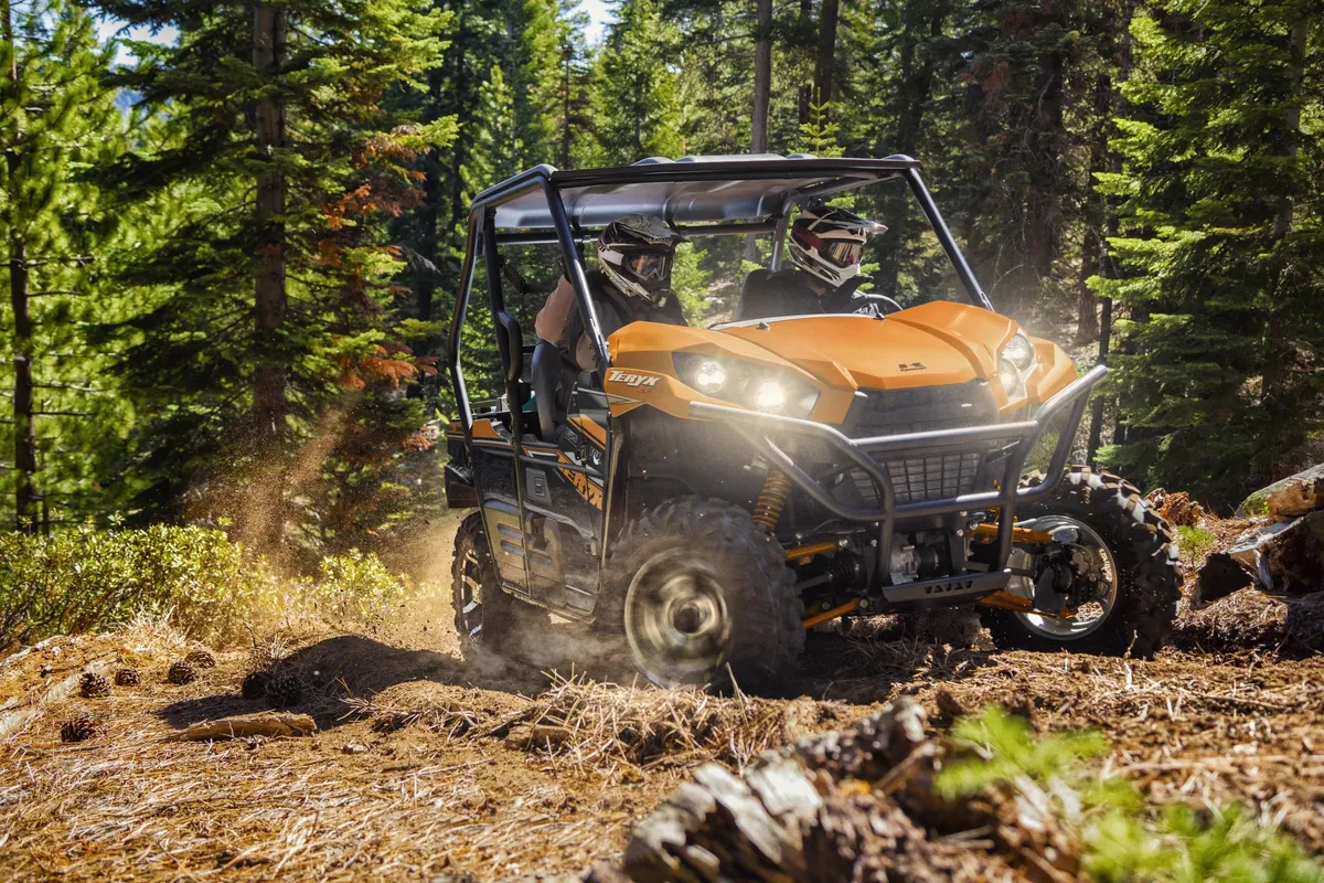 A dynamic outdoor shot featuring an ATV with two riders navigating a dense forest trail. Sunlight filters through tall trees, suggesting an exciting off-road experience in a natural mountain setting.