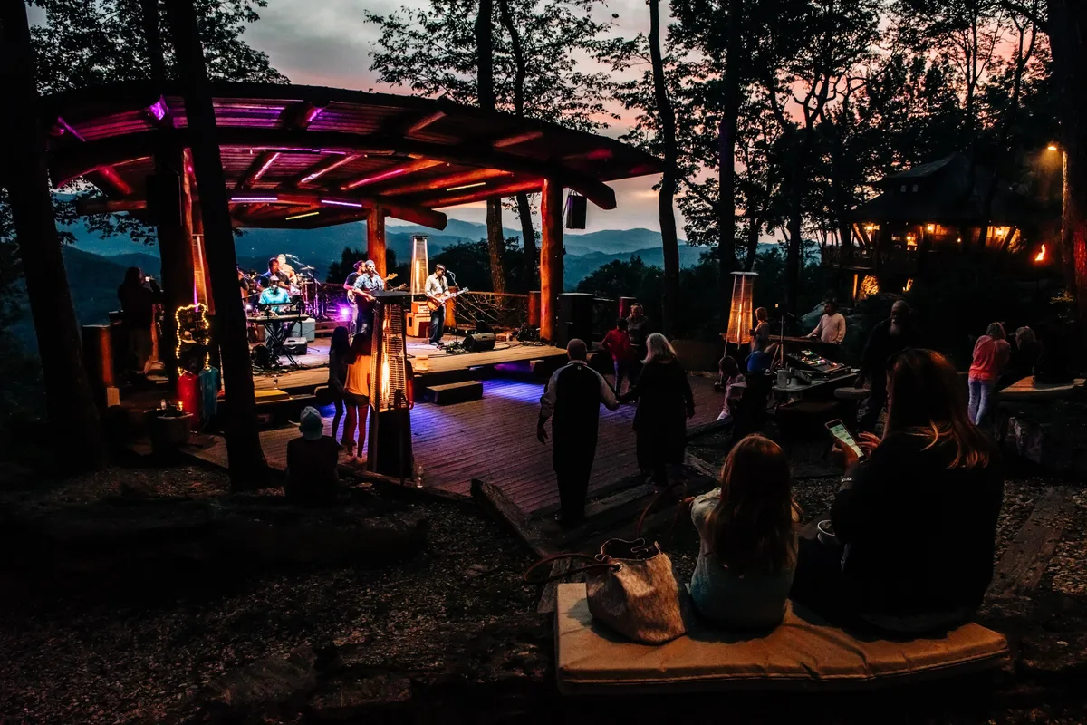 A lively outdoor concert scene featuring musicians on stage, an engaged audience, and a stunning mountain backdrop. The atmosphere is joyful and communal, emphasizing shared experiences and enjoyment of live music in a natural setting.