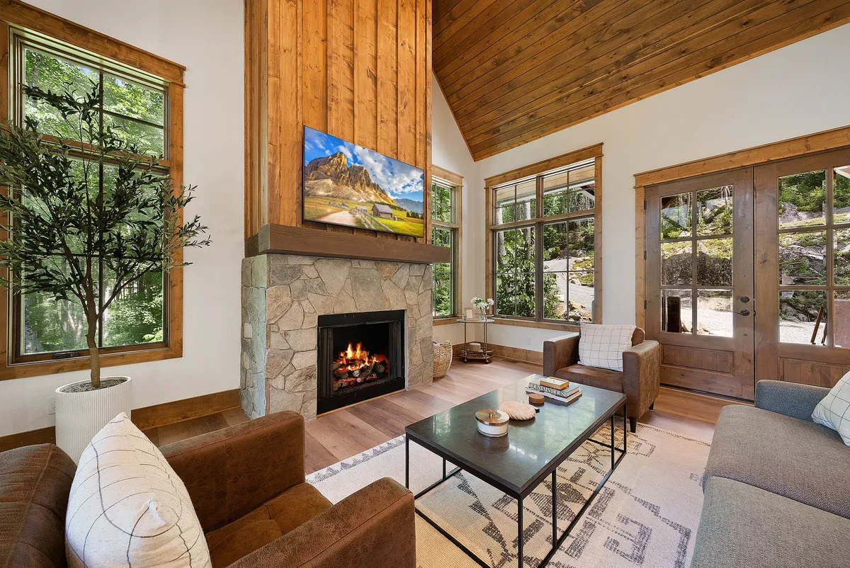 A spacious and inviting living area featuring a grand stone fireplace, large windows that bathe the room in natural light, exposed wooden beams, and a television. Plush sofas are arranged around the hearth, creating a cozy yet open space perfect for relaxing, socializing, or enjoying a movie night.