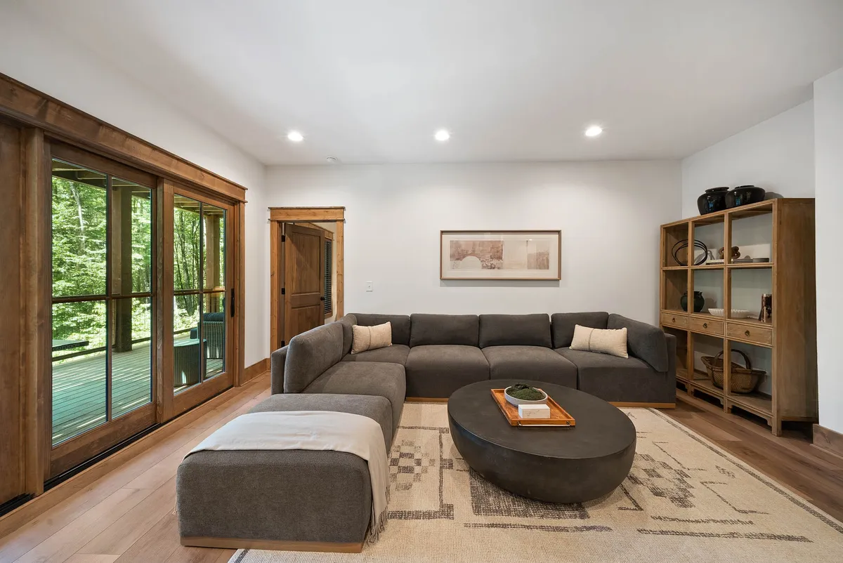 A cozy lounge area with a large sectional sofa, coffee table, bookshelf, and broad windows letting in natural light. The space is ideal for relaxing, reading, or gathering for games and conversation.